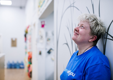 A women with short, curly blond hair and a blue t-shirt smiling, seated in a brightly lit room with wall designs and colorful items in the background.
