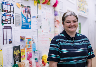 A smiling woman in a striped polo shirt stands in front of a vibrant community bulletin board adorned with various colorful notices and decorations.