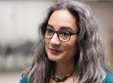 A woman with shoulder-length gray hair and glasses, wearing a teal top and black cardigan, in an office setting.