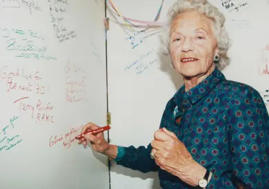 An elderly woman with curly gray hair, wearing a blue patterned blouse, smiles as she writes on a whiteboard holding a red marker.