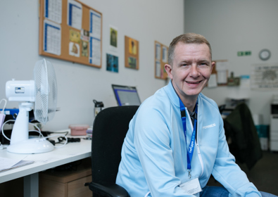 A smiling man wearing a blue shirt and a lanyard sits at a office desk with a small white fan, surrounded by various items and pictures on the wall.