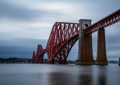 Shot of bridge, a cantilever railway bridge over a body of water under cloudy skies, highlighting its red colour and massive stone piers.