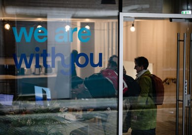 Office door with the words "we are with you" printed on it, visible indoor office setup with a person working on a laptop.