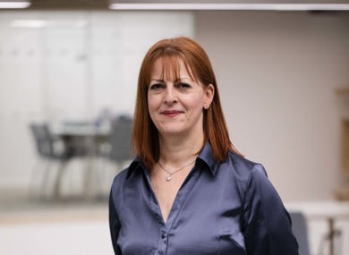 A woman with shoulder-length red hair, wearing a navy blue blouse and black trousers, stands in an office with a whiteboard in the background.